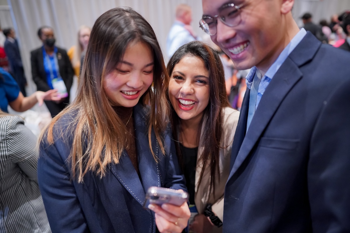 three professionally dressed people looking at an iPhone and laughing